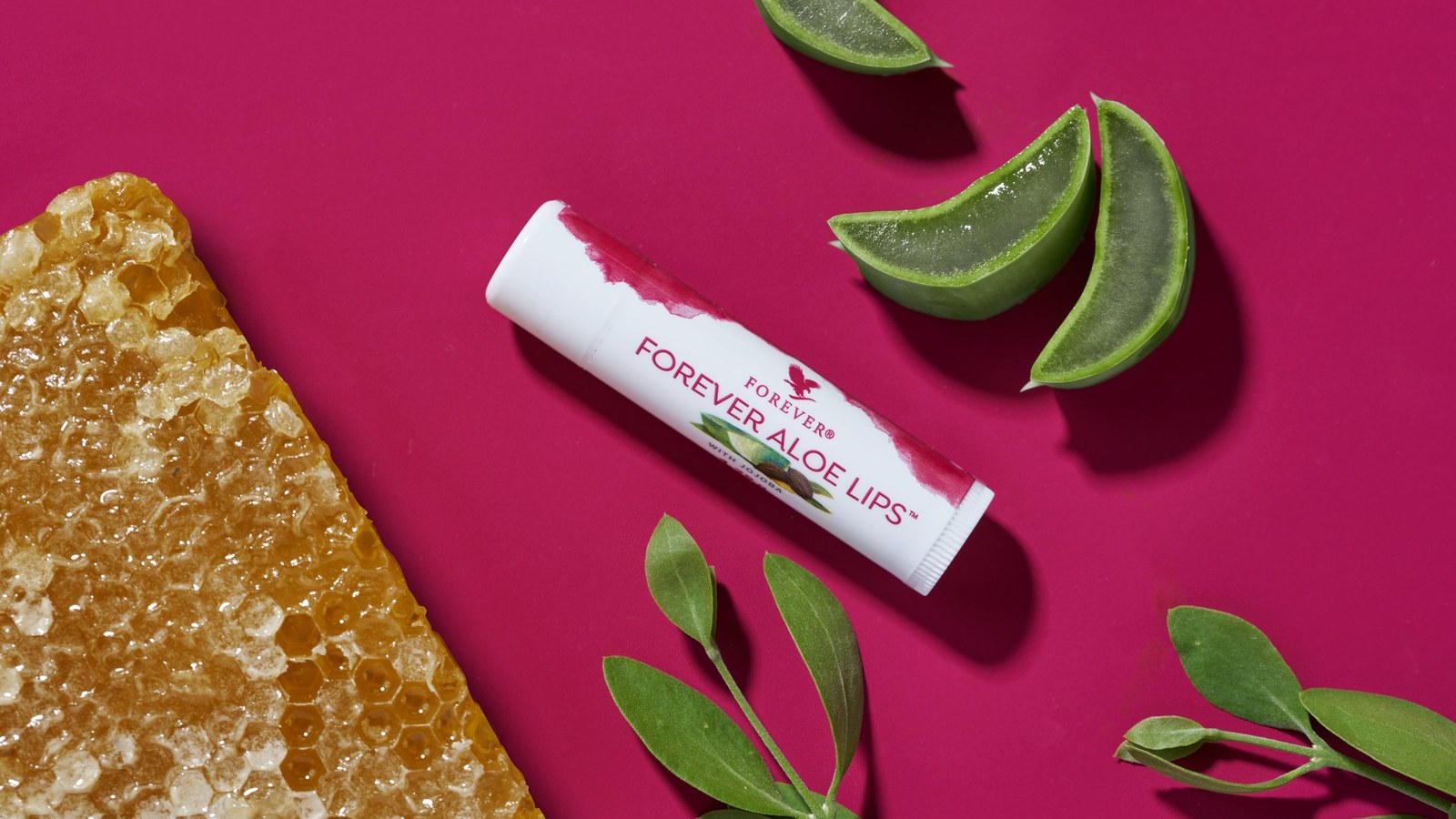 Forever Aloe Lips, Aloe-Vera-Lippenpflegestift, mit Aloe-Vera-Scheiben und Wabe auf magentafarbenem Hintergrund